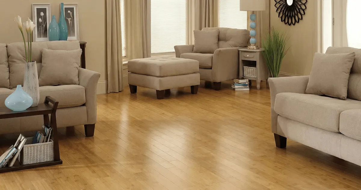 A living room with professionally installed bamboo flooring, beige sofas, a matching ottoman, a side table with a blue lamp, vases, framed pictures, and large windows with blinds and curtains in Columbus, OH
